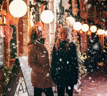 Two women pictured in Belleville's Downtown District during Enchanted. Light snow falling and christmas lights can be seen.