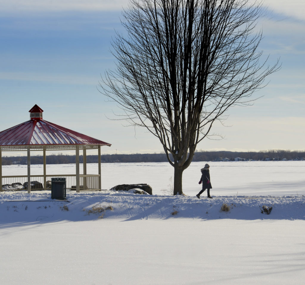 A person walks along the waterfront trail past a red gazebo on a sunny winter day. The bay is frozen and covered in snow.