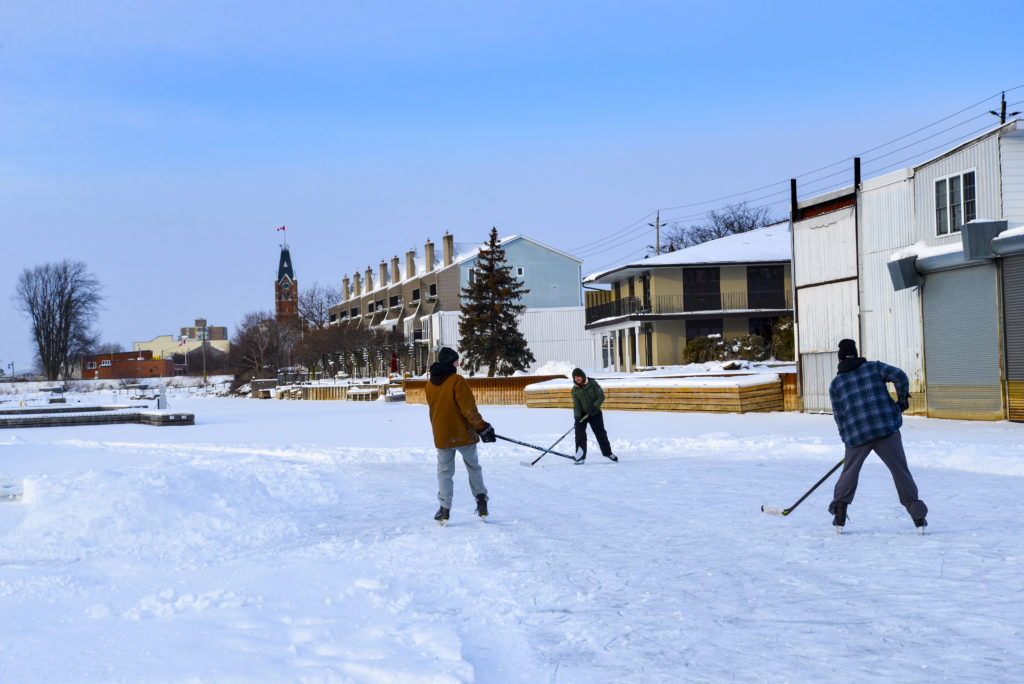 Three people skating and playing hockey on the ice in Victoria Harbour. Belleville City Hall is visible in the background.