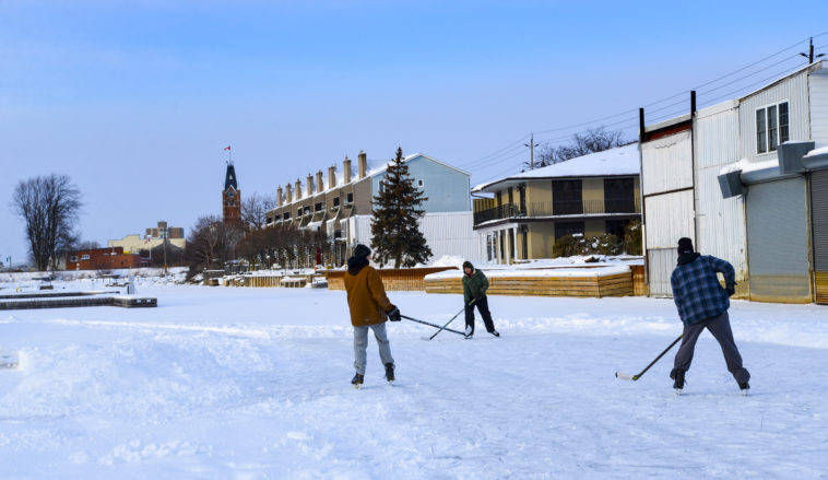 Three people skating and playing hockey on the ice in Victoria Harbour. Belleville City Hall is visible in the background.